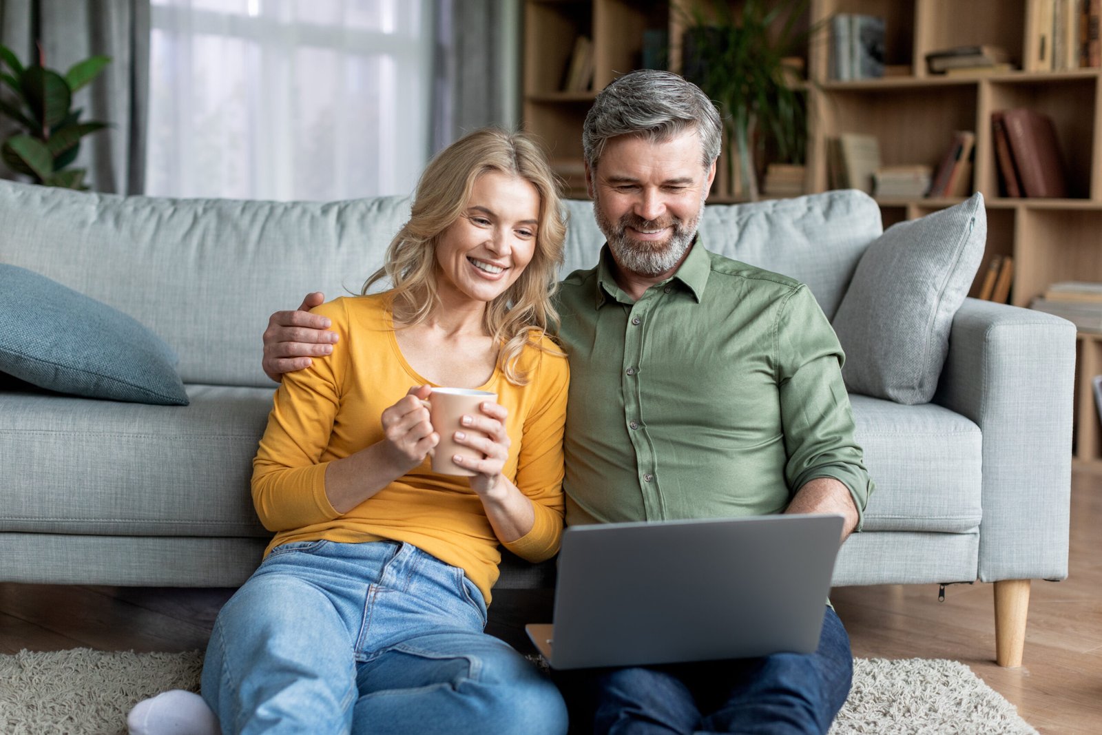 Happy Middle Aged Couple Spending Time With Laptop At Home, Smiling Mature Wife And Husband Sitting On Floor And Looking At Computer Screen, Watching Movies Together, Woman Drinking Coffee