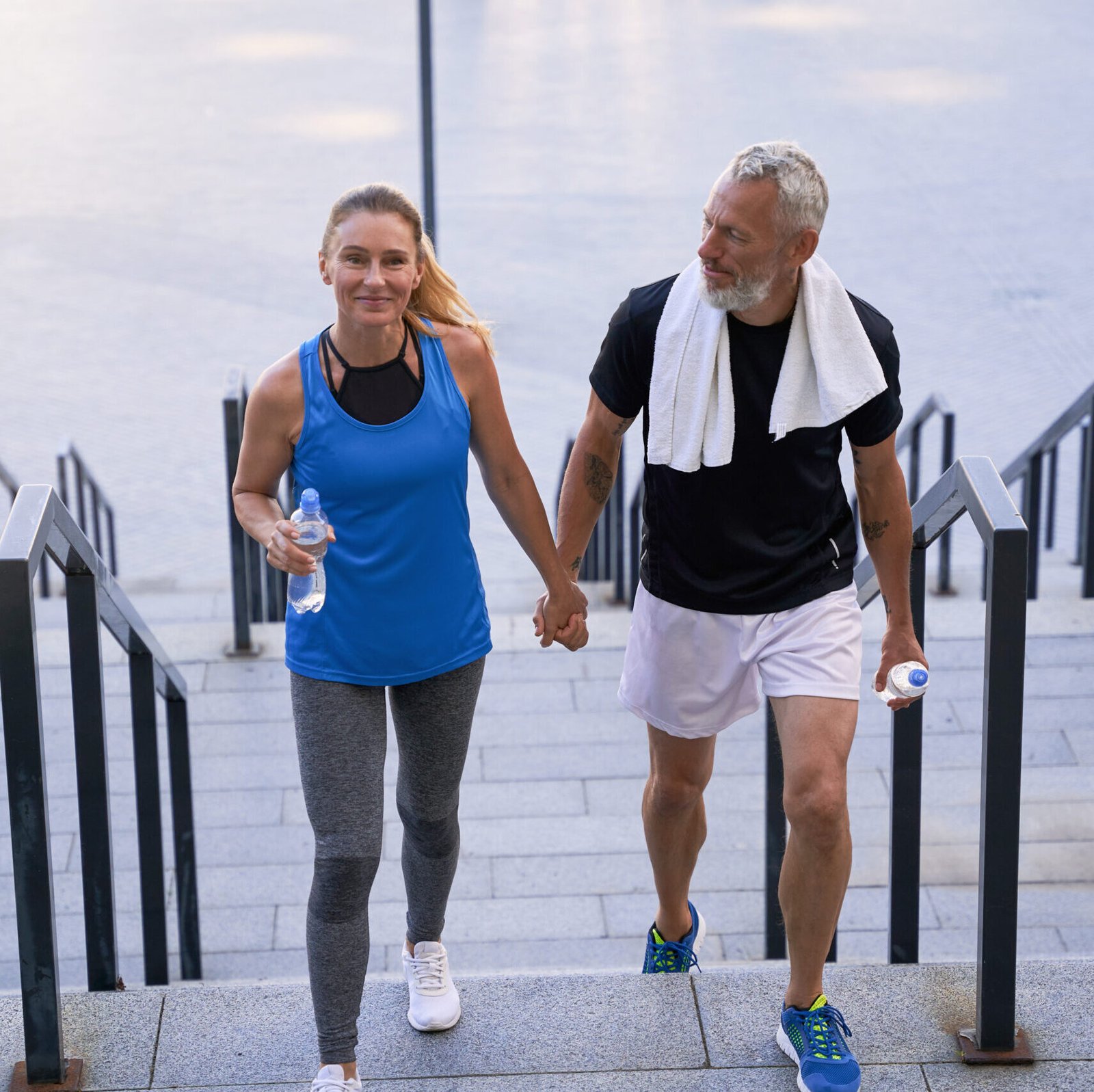 Eternal Health Solution - Sporty middle aged man and woman in sportswear looking happy, holding hands while walking up the stairs after training together in the city